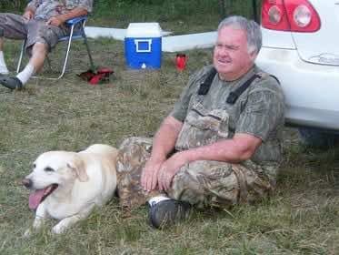 Top Dogs photo for Boo: a yellow Labrador lies on the grass beside a man in camouflage sitting on the ground; another person sits in a camp chair behind them.