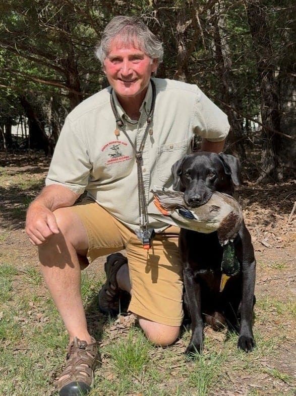 Top Dogs photo for Bracken: a man in a khaki shirt and shorts kneels beside a black Labrador holding a duck.