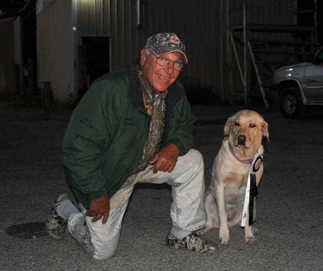 Top Dogs photo for Chance: a man in a green jacket and cap kneels beside a sitting yellow Labrador, outdoors after dark.