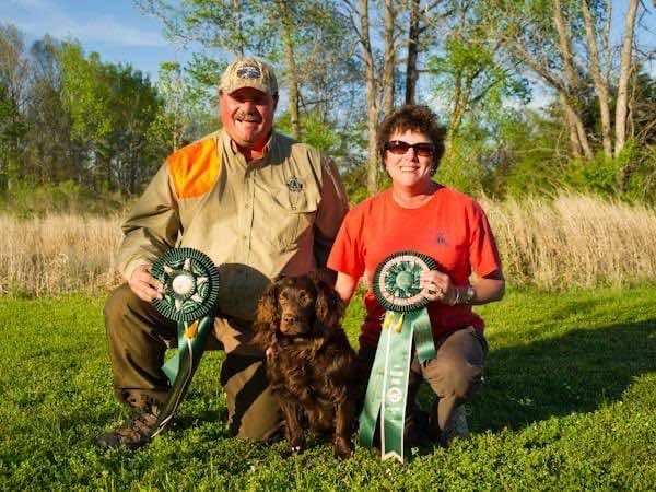 Top Dogs photo for Chief: a man and a woman kneel on the grass with a brown spaniel between them, each holding a green rosette ribbon.