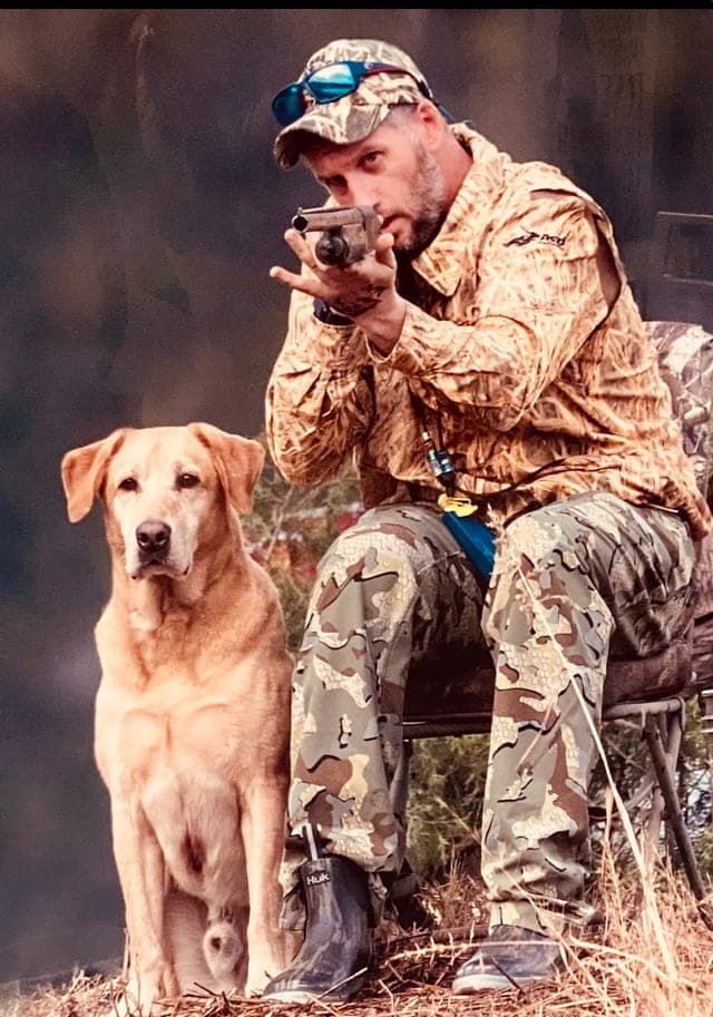Top Dogs photo for Gibbs: a seated man in camouflage aims a shotgun toward the camera beside a sitting yellow Labrador, in front of a studio backdrop.
