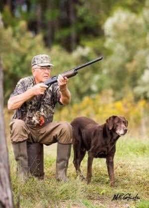 Top Dogs photo for Jimbo: a chocolate Labrador stands beside a seated man in camouflage holding a shotgun, at the edge of a field.